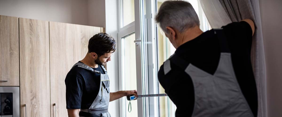 Two professional workers in uniform using tape measure while measuring window for installing blinds indoors. Construction and maintenance concept. Focus on young man. Horizontal shot